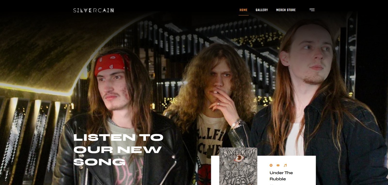 Silvercain home page: the three-piece photographed at night under the line Listen To Our New Song, with a player for Under The Rubble